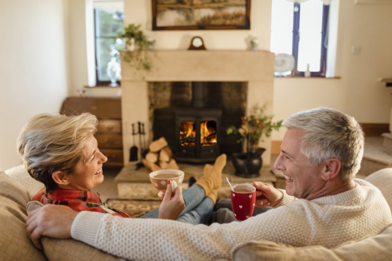How to Keep Warm During Extreme Cold Weather. Mature couple sat in front of a fire drinking hot chocolate from mugs.
